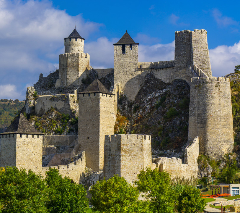 Historic Golubac Fortress stands proudly over the serene Danube River in Serbia, surrounded by lush greenery and dramatic clouds against a bright blue sky