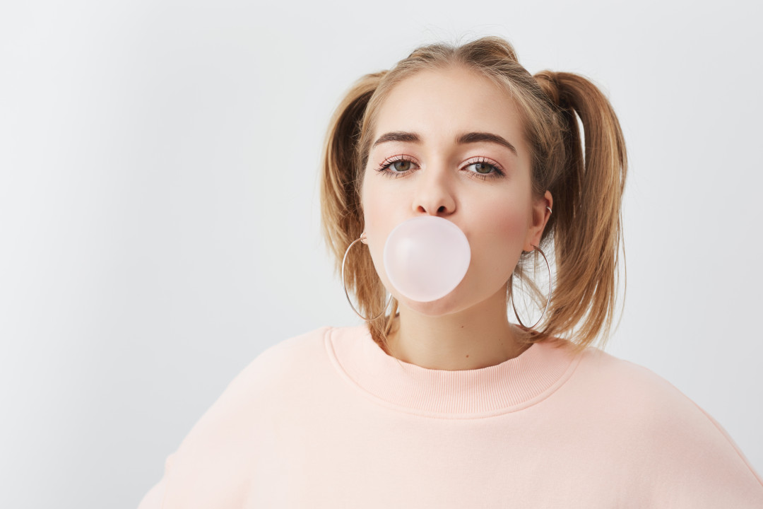 Beautiful, pretty and funny young girl blowing bubble from chewing gum. Blonde female with two ponytails on head wearing pink sweatshirt posing against gray background.